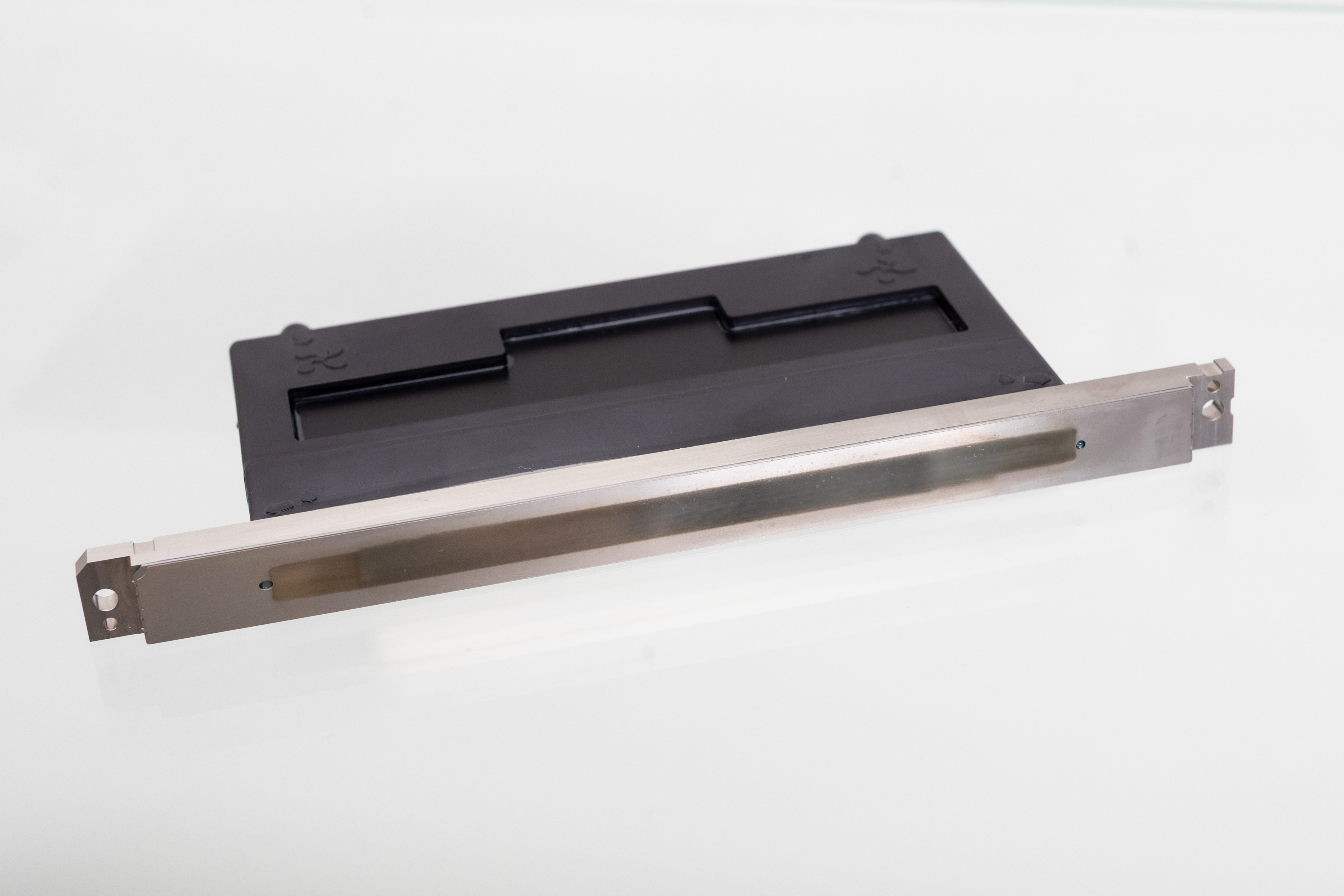 Photograph of a black and silver electronic component, likely a card reader or slot interface, placed on a white surface. The component features a rectangular black plastic housing with a horizontal slot and a silver metallic front panel with mounting holes on each end.