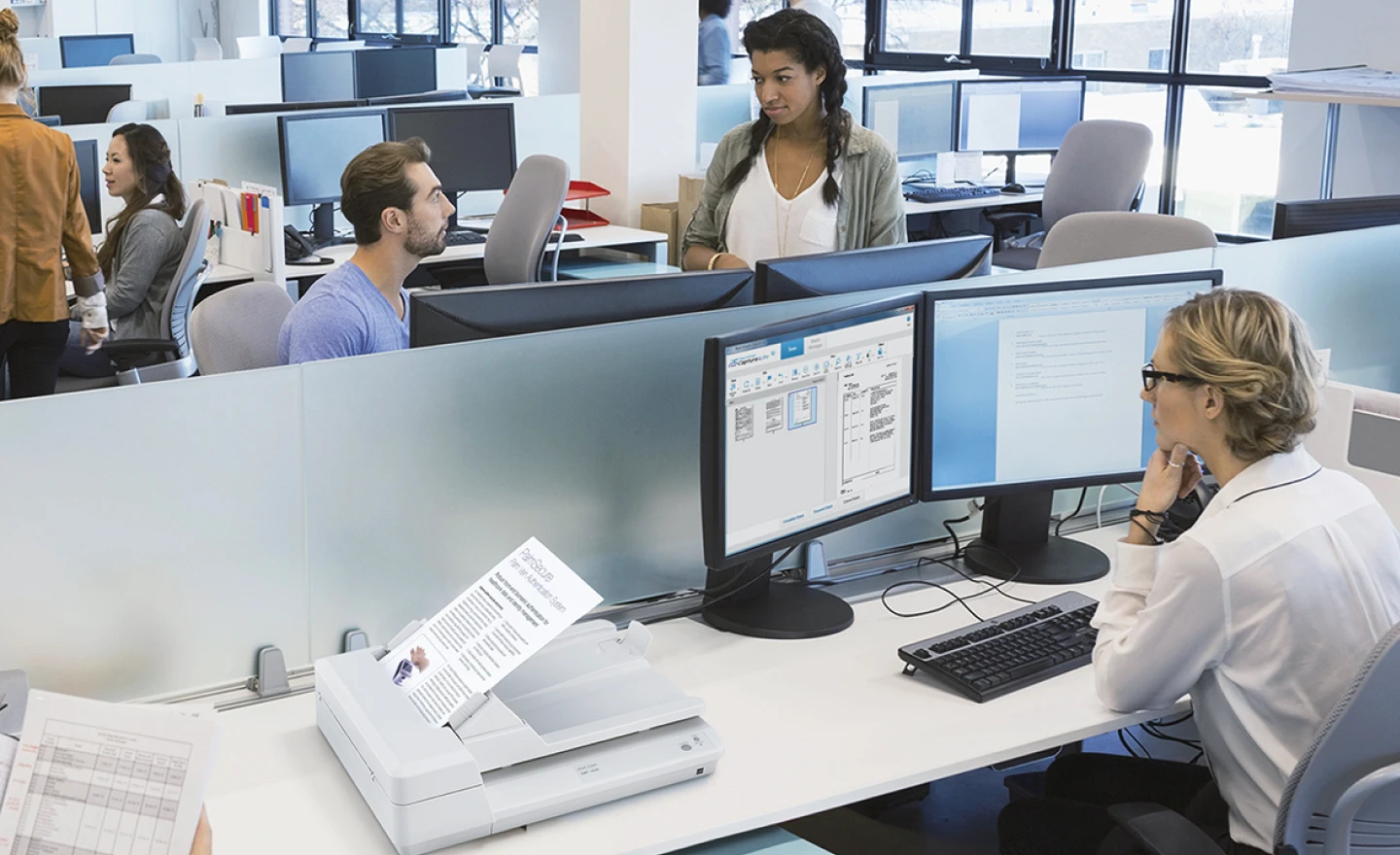 An office environment with several people working in cubicles. In the foreground, a woman with glasses sits at a desk, looking at two computer monitors. A document scanner is in front of her, actively scanning a paper. Behind her, a man in a purple shirt looks up at a woman with dark curly hair who is standing and looking at him over a cubicle wall. Other office workers and computers are visible in the background.