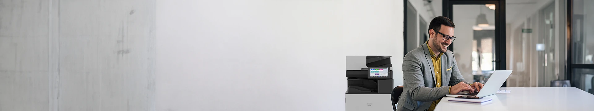  A man in glasses and a grey suit jacket sits at a white table, smiling as he types on a laptop. To his left, a Ricoh printer is visible. The background is a bright office space.